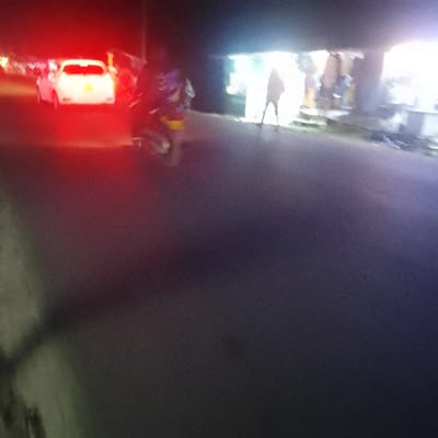 At night, a brightly lit roadside shop stands next to a dark street. A person is silhouetted under the shop's awning. Further down the road, a motorcycle moves past a stopped car, its brake lights glowing red. The image is grainy, particularly in the foreground where the road is in focus.
