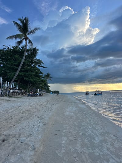 The image depicts a beach scene in Baan Thong Po, Thailand, during what appears to be late afternoon or early evening. The sky is dramatic, with a mix of dark, heavy clouds on the left, backlit by the golden glow of the setting sun on the right. This contrast creates a dynamic and moody atmosphere.
The foreground is dominated by a sandy beach, showing footprints, suggesting recent activity. The ocean stretches out to the horizon, with its surface rippling and reflecting the light. A few small boats, possibly fishing vessels, are anchored in the water.
Along the edge of the beach, near the tree line, there's a set of outdoor seating areas, with tables and chairs, and umbrellas, indicating a restaurant or bar. Several people are visible seated at these tables, enjoying the view and the cooling evening air. Lush green trees, including palm trees, line the back of the beach, adding to the tropical setting. One prominent palm tree leans diagonally across the frame.
The overall impression is one of a peaceful evening on a tropical coast, with the natural beauty of the sky and sea complemented by the human presence of the beachside establishment. The weather appears to be transitioning, with the possibility of rain hinted at by the dark clouds.