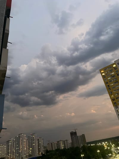 The image depicts a view of the city of Brovary, Ukraine, during twilight. The sky is filled with dramatic, dark clouds, suggesting an approaching storm or the aftermath of one. In the foreground and mid-ground, numerous high-rise apartment buildings are visible, many of which have illuminated windows, indicating that residents are inside. One distinct yellow building on the right side of the frame is particularly prominent. Further into the distance, more buildings and a construction crane can be observed, suggesting ongoing development in the city. The lower portion of the image shows trees and streetlights, illuminating what appears to be a parking area with several vehicles. The overall impression is one of a residential urban landscape under a moody, atmospheric sky. No people are clearly visible in the frame, and no specific actions or interactions are depicted.