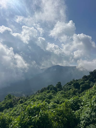 The image displays a sprawling, verdant mountainous landscape under a dynamic sky in the vicinity of El Congo, El Salvador. The upper approximately 40% of the vertical frame is occupied by a light blue sky with numerous white and grey clouds. Distinct sunrays are visible emanating from the upper-left, piercing through clouds. The clouds vary in form from fluffy cumulus to more layered, darker formations, creating a sense of atmospheric depth.

Below the sky and clouds, a series of mountains or large hills extend across the mid-ground. The closest slopes, filling the lower 60% of the frame, are densely covered with vibrant, varied green foliage, characteristic of a lush forest or extensive vegetation on a steep incline. Individual leaves and clusters of plant life are clearly distinguishable, with areas of both bright illumination and shadowed depth.

Further in the distance, a larger mountain range is partially enshrouded by mist and lower clouds, rendering its contours softer and more muted in tone. The highest visible peak of this range appears broad and somewhat flattened against the horizon line. In the mid-ground, between the foreground foliage and the distant mountains, faint indications of human settlement are visible; specifically, several small structures, possibly with reddish roofs, can be discerned within a distant valley or lower area. No individuals are present or engaged in any actions within the frame. The scene is illuminated by natural daylight, contributing to the overall bright and clear atmospheric conditions despite the significant cloud cover and atmospheric haze around the distant peaks.