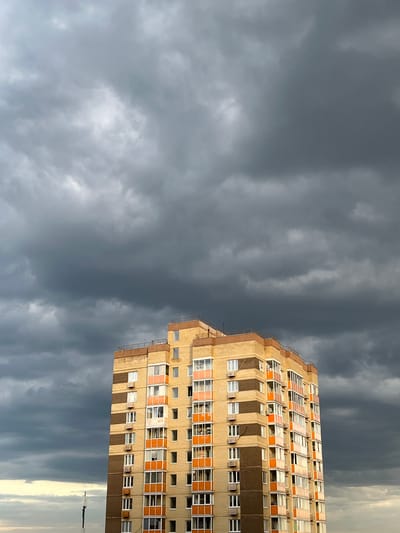 This is a low-angle, full shot of a modern apartment building in Lyubertsy, Russia, set against a dramatic backdrop of dark, stormy clouds. The building is tall and rectangular, constructed with light-colored bricks and accented with horizontal bands of darker brown brickwork. Each floor features multiple windows and enclosed balconies with orange trim. The building is illuminated by natural light that highlights its facade, while the sky above is filled with ominous, grey storm clouds, suggesting an approaching downpour. The lower portion of the image shows a sliver of the horizon with a faint glow of light, possibly from the sun breaking through elsewhere. A single antenna-like structure is visible in the lower left corner. There are no people or active events depicted in the scene.