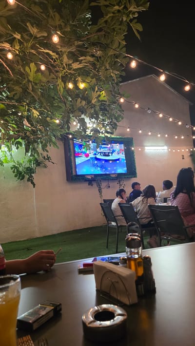 The image depicts an outdoor seating area at night, likely a restaurant or cafe, decorated with string lights and foliage. A large television screen mounted on a wall displays a program. Several patrons are seated at tables, some interacting while others appear to be watching the screen. On a table in the foreground, a glass of orange juice, a pack of cigarettes, an ashtray, and a condiment set are visible, suggesting a relaxed dining or social setting. The overall ambiance is casual and inviting, enhanced by the warm glow of the lights.