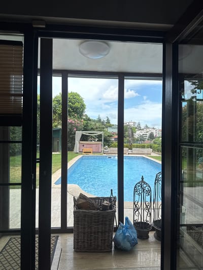 The image shows a view through an open doorway onto an outdoor scene. A bright blue swimming pool dominates the right side of the frame, with a tiled edge and a light-colored patio surrounding it. Beyond the pool, there is a lush green lawn and trees. In the distance, several white buildings are visible on a hillside, suggesting an urban or suburban setting.

On the patio, several objects are present. A large wicker basket filled with logs sits near the pool's edge. Further to the right, a blue plastic bag is tied shut, and next to it are two ornate black metal stands, possibly decorative planters or candle holders. A small pink gazebo-like structure with white curtains is visible on the lawn, adding a touch of whimsical design to the outdoor space. The sky is blue with scattered white clouds, indicating fair weather.

The perspective suggests the photo was taken from inside a building, looking out through a large window or door frame. The interior visible in the foreground is dimly lit and features dark-colored framing. The overall impression is of a pleasant, sunny day at a residential or resort-like location. The image does not contain any visible text. The location context provided, Beşiktaş, Turkey, aligns with the Mediterranean architectural style of the distant buildings and the lush greenery.
