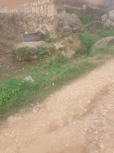 An outdoor, eye-level, medium shot captures a scene along Rukuba Road, Nigeria. The foreground is dominated by a dirt road with scattered small stones and tire tracks. To the left, a patch of lush green grass grows, interspersed with a few small pieces of litter, including what appears to be a white plastic bag and some colorful wrappers. In the middle ground, a weathered, rusty metal barrel, with a blue band around its base, sits atop a stone structure. Behind the barrel, a rocky embankment rises, partially covered with more greenery and larger stones. A weathered stone wall forms the background, with remnants of what appears to be a building. The overall impression is one of a rustic and somewhat neglected environment.
