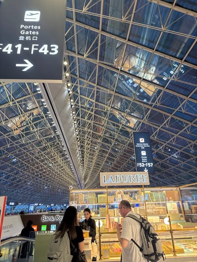 The image displays an interior view of a modern airport terminal at Paris-Charles de Gaulle Airport in Le Mesnil-Amelot, France. A distinctive high, arched ceiling composed of a metal grid and glass panels dominates the upper portion of the frame. Below, three individuals are engaged at a Ladurée confectionery counter. A female, seen from behind with dark hair and a grey backpack, stands on the left. To her right, a male with short hair, glasses, and a white t-shirt, wearing a grey backpack, faces the counter and looks towards the employee. The employee, a female with dark hair in a dark uniform, is positioned behind the glass display case, which holds various pastel-colored boxed sweets and gift sets. Overhead, two black directional signs are visible: one indicating "Portes / Gates / 登机口 F41-F43" with a right-pointing arrow, and another further right showing "Service clients / Customer Service / 旅客服务" and "Portes / Gates / 登机口 F52-F57". In the background to the left, a down-escalator is visible, along with distant signage for "Bars, Rest..." and a Starbucks logo, indicating other commercial areas within the terminal. The environment is brightly lit, with numerous lights integrated into the ceiling structure.