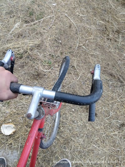 The image depicts a first-person perspective, looking down at a red bicycle being held by a single visible hand in Balgarsko Slivovo, Bulgaria, on March 28, 2026, at 16:07. A light-skinned human hand grips the worn black rubberized left drop handlebar of the bicycle. The bicycle features a red frame with visible wear and scratches, silver-colored drop handlebars, and silver brake levers with red buttons. The ground beneath consists primarily of dry, light brown straw-like grass and dead vegetation, with minimal green patches. A piece of white, irregular bread lies on the ground to the left of the bicycle's front wheel. The tips of dark-colored shoes are visible at the bottom edges of the frame.
