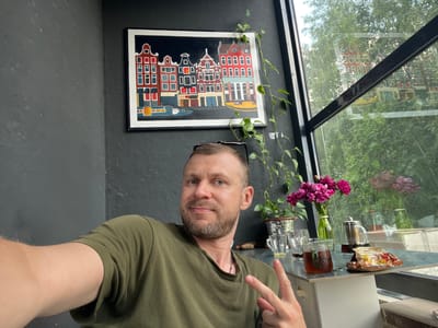 The image shows a man taking a selfie in an indoor setting. The man, with short brown hair and a beard, is wearing a green t-shirt and making a peace sign with his right hand. He appears to be smiling slightly.
On a glass-topped table in front of him, there are several objects: a package of what looks like biscuits, a glass mug with a brown tint, a small metal pitcher, two clear glass cups, and a green vase with pink flowers. A green vine with leaves drapes from the ceiling or a nearby shelf. Through the large window behind the table, lush green trees and foliage are visible, suggesting a pleasant outdoor environment, possibly a sunny day.
On the dark gray wall behind the man, a framed painting depicts a row of colorful, narrow buildings, characteristic of Dutch architecture. The overall scene suggests a relaxed moment, perhaps during a coffee break or a visit to a cafe. The lighting indicates it is daytime. There is no visible text in the image.