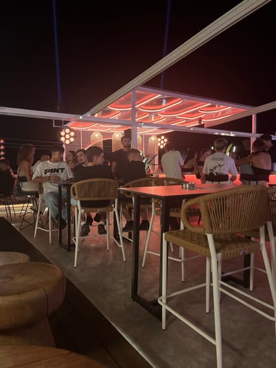 This image depicts a lively evening scene at a rooftop bar or club on Ofrynio Beach, Greece. The setting is characterized by modern, open-air architecture with a white roof structure illuminated by vibrant red neon lights. Several groups of people are seated at high-top tables, engaged in conversation and enjoying the atmosphere.
In the foreground, a man in a white t-shirt with the word "PINT" printed on it sits at a black high-top table, looking down at his phone. He is wearing blue jeans. To his right, another man in a black t-shirt is seated. The chairs are woven rattan with white metal legs, creating a stylish, contemporary look.
Further into the scene, a diverse crowd is visible. Some individuals are standing, while others are seated, their faces dimly lit by the ambient red glow. Behind the patrons, decorative rattan lamps hang from the ceiling, adding to the bohemian-chic ambiance. The floor appears to be a polished concrete or stone surface, reflecting the colorful lighting.
In the background, a bright blue light beam cuts through the dark night sky, suggesting a powerful spotlight or laser effect, further enhancing the club-like atmosphere. The overall impression is one of a popular nightspot, filled with young adults enjoying a night out by the sea.