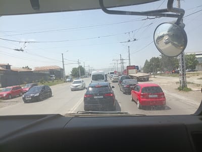 The image shows a street scene in Osh City, Kyrgyzstan, viewed from the perspective of a vehicle driver. The scene is characterized by moderate traffic congestion on a sunny day. Multiple vehicles are visible on the road, including cars, vans, and trucks.

In the foreground, the dashboard of the vehicle is visible, with a large rearview mirror mounted on the driver's side. The mirror reflects a portion of the road and the traffic behind.

The road appears to be a multi-lane asphalt surface. Several cars are stopped or moving slowly in traffic. Notable vehicles include a black BMW SUV with license plate 01 619 AZH, a red hatchback with license plate 06 258 ALD, and a white sedan with license plate 01 263 DHN. Other cars in various colors are also present.

On the sides of the road, there are buildings with corrugated metal roofs, trees, and utility poles with overhead wires, indicative of an urban or semi-urban environment. The sky is clear and blue.

The overall impression is a typical traffic situation in a city, with vehicles moving along a road under a bright sky. There are no people clearly visible in the image.