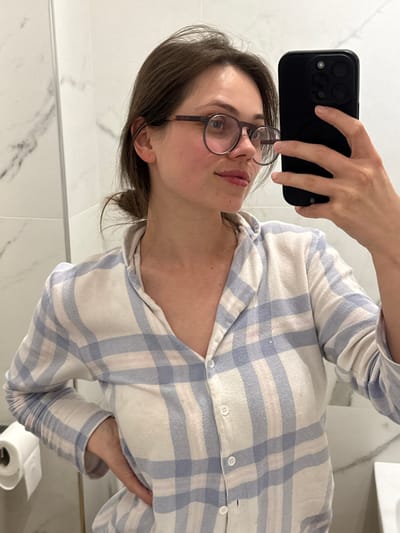 The image depicts a female individual taking a mirror selfie within an interior setting, likely a bathroom, as indicated by white marble-patterned wall tiles and a visible toilet paper roll on the left. The subject, appearing to be in her twenties or thirties, has light skin, dark brown hair styled in a bun, and wears round, dark-framed eyeglasses. She is dressed in a long-sleeved, collared shirt featuring a light blue and white plaid pattern, unbuttoned at the neckline. Her left hand holds a black smartphone, equipped with a triple-lens camera system, angled to capture her reflection. Her right hand is placed on her hip. She maintains a slight smile while looking towards her reflection. The specific location within the city of Scafati, Italy, cannot be determined from the visual information provided.