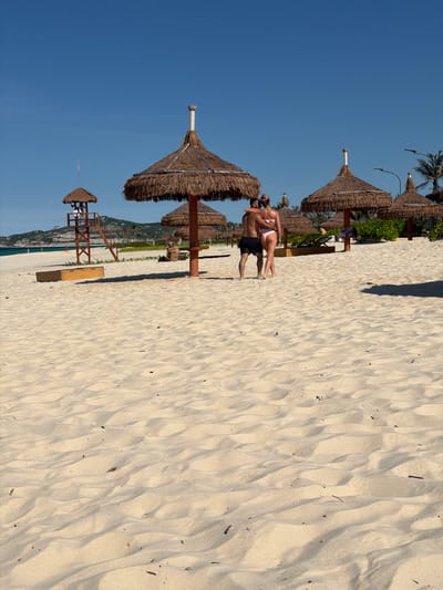 On a wide, sandy beach in Bac Cam Ranh Ward, Vietnam, a couple stands embracing near a cluster of thatched umbrellas. The man, wearing black swim shorts, has his arm around the woman, who is in a pink bikini. Behind them, several straw-covered beach umbrellas stand in a line, with some beach chairs visible further back. A lifeguard tower with a person in it is positioned to the left, and the turquoise ocean stretches out towards a distant, green-covered hill under a clear blue sky. The foreground of the image is dominated by the texture of the sand, showing ripples and a few scattered pieces of debris.