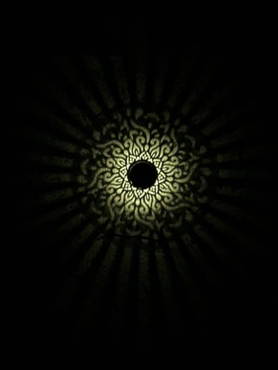 The image displays a dark, textured surface illuminated by a central light source. At the core is a distinct dark circular void, from which a brightly illuminated, intricate, symmetrical pattern resembling a mandala or ornate scrollwork radiates outward. The pattern is rendered in a warm, pale yellow or light beige hue against the darker backdrop. Extending from this central illuminated design are prominent, straight, dark radial shadows, creating a starburst effect. The surrounding surface exhibits a rough, granular texture, suggestive of concrete or stucco. No individuals, explicit actions, or specific contextual identifiers for a geographical location are discernible within the frame.