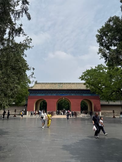 The image shows a red and tan traditional Chinese gate with three arched doorways, likely located in the Dongcheng District of China. The gate features a tiled roof and is surrounded by trees. People are seen walking in front of the gate, some of whom are wearing bright clothing. The environment is a paved area with trees visible in the background, under a cloudy sky.