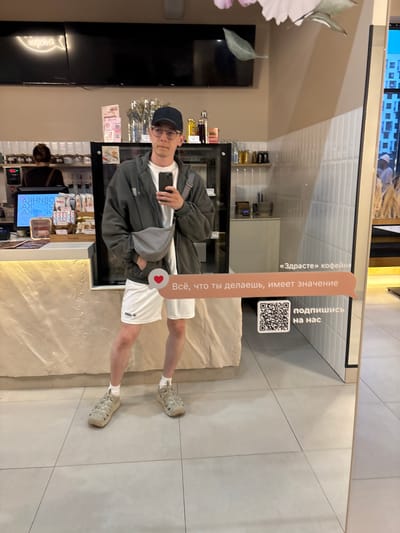 A young man wearing a grey hooded jacket, white t-shirt, white shorts, a black baseball cap, glasses, and beige sandals is taking a mirror selfie. He is holding a black smartphone in his right hand. He has a grey fanny pack worn across his chest. The man is standing in what appears to be a coffee shop or cafe. The background shows shelves stocked with various items, a digital menu board, and a tiled wall. On the mirror, there are superimposed text bubbles with Russian lettering and a QR code. The text includes "«Здрасте» кофейня", "Всё, что ты делаешь, имеет значение", and "подпишись на нас". The location is identified as the city of Misaylovo, Russia, based on information from the superimposed text.