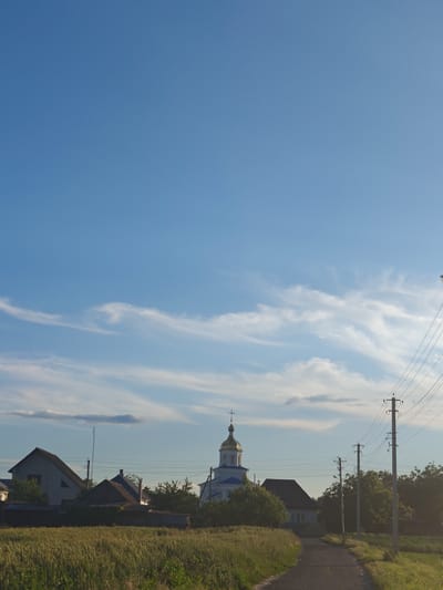 The image depicts a rural landscape in Kotelva, Ukraine, bathed in the warm light of a clear day with scattered white clouds. In the foreground, a field of wheat or similar tall grass extends across the bottom of the frame. A dirt or paved road curves gently from the bottom right corner towards the center of the image, disappearing behind vegetation.
On the left side of the road, several residential buildings are visible, characterized by their angular roofs and muted colors. Behind these homes, trees form a dense green backdrop. Dominating the mid-ground and slightly to the left of the road is a white church with a golden dome and a cross at its apex. The church is a focal point, its architecture standing out against the clear blue sky.
To the right of the road, a series of utility poles with numerous wires stretch into the distance, indicating the presence of electrical infrastructure. More trees and vegetation are visible behind the poles. The overall scene conveys a sense of peaceful, rural life in a Ukrainian village under a vast, serene sky. No people are visible in the image.