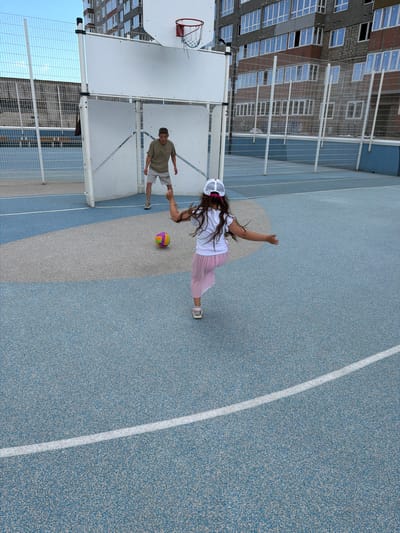 The image depicts a children's sports ground in Novorossiysk, Russia, likely on a sunny day with a clear sky. In the foreground, a young girl with long brown hair, wearing a white t-shirt, a pink pleated skirt, a white baseball cap with a pink brim, and white shoes, is positioned on the blue and gray-patterned asphalt court. She is captured mid-motion, facing away from the camera, with her right arm extended and her left arm bent, suggesting she is either kicking or about to kick a colorful volleyball. The volleyball, featuring segments of yellow, orange, purple, and green, rests on the court in front of her. In the background, a young boy, dressed in a khaki t-shirt and light gray shorts, stands in front of a white sports structure with a basketball hoop. The structure appears to be a wall or enclosure used for sports. The boy is looking towards the girl. Behind the sports structure, a tall, multi-story building with numerous windows is visible, indicating an urban setting. A metal fence surrounds the sports area, and further in the distance, another building and a fence can be seen. The overall scene is one of children engaged in outdoor recreational activity.