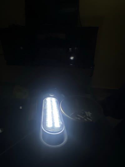 A brightly illuminated rectangular LED light fixture is affixed to a dark surface. Above the LED fixture is a circular object. To the left of the light, a darker rectangular shape is discernible against a dark backdrop. No people are visible. The environment appears to be indoors, with limited ambient light. No location can be found within the city of Hanwa, Nigeria.