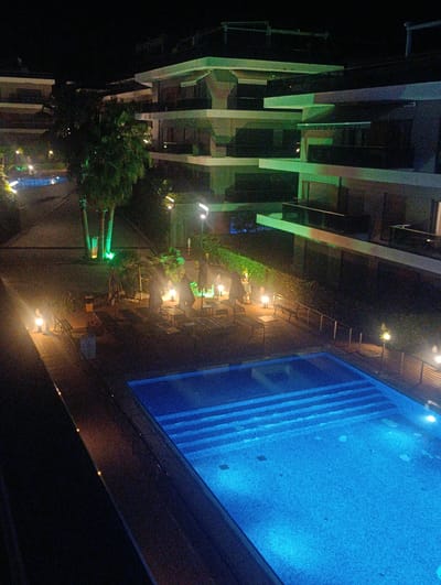 An aerial, nighttime view shows a resort complex in Urla, Turkey, featuring a brightly lit swimming pool. The pool, tiled in a mosaic pattern, glows with blue underwater lights, and steps descend into its depths. Surrounding the pool is a wooden deck with lounge chairs, umbrellas, and low-lying lamps casting a warm glow.

Behind the pool area, trees with illuminated trunks stand tall. In the background, a multi-story building with modern architecture and green accent lighting is visible. The building features balconies and multiple levels, suggesting it is a hotel or apartment complex. The overall scene depicts a serene and inviting atmosphere, perfect for a nighttime relaxation or gathering.