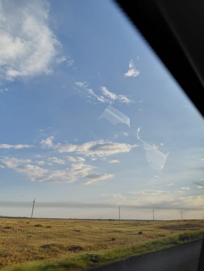 The image depicts a vast, dry, golden-brown grassy field under a pale blue sky filled with scattered white clouds. The horizon is flat and stretches across the scene, punctuated by a few distant utility poles with wires. In the foreground, a dark, blurred surface, possibly a road or pathway, runs diagonally across the bottom of the frame. The image appears to be taken from inside a vehicle, as evidenced by the reflection of the vehicle's interior on the window, which includes the top of a car seat and a portion of the windshield frame. The light suggests it is either morning or late afternoon. The location is described as Molodezhny, Kazakhstan.