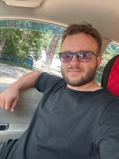 A man is sitting in a car in Blagoveshchensk, Russia, with a black t-shirt on and sunglasses. The man has a beard and short brown hair. He is resting his left arm on the car door. Outside the car window, there are trees and buildings visible, indicating an urban or suburban setting. The interior of the car is also visible, with a beige seat and headliner. The sun is shining, casting shadows and highlighting the textures of the car's interior and the man's skin.