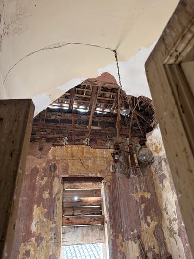 The image presents the interior of a severely dilapidated room within a building located in Catania, Italy. The perspective is a low-angle shot, looking upwards towards the ceiling and walls.
The arched ceiling, originally plastered, exhibits extensive damage, with large sections having collapsed. Exposed wooden roof beams and lathing are visible through the gaps, with bright sunlight penetrating from above, suggesting a partial roof collapse or missing roof tiles. The remaining plaster on the ceiling is light cream-colored, showing significant cracking, water damage, and peeling paint. A dark, thin cable runs along a crack in the ceiling.
Below the ceiling, the walls are in an advanced state of decay. Multiple layers of paint, primarily faded reddish-brown and yellow-ochre over lighter undercoats, are extensively peeling and flaking. Higher portions of the walls reveal rough, dark-colored exposed brickwork where plaster has completely deteriorated.
A prominent, aged metal chandelier, likely rusted, is suspended by a chain from a point of damage in the ceiling, slightly right of center. The chandelier features ornate, possibly scrollwork, elements and contains two visible clear glass light bulbs. To the left and slightly higher than the main chandelier, a simpler light fixture, appearing as a bare bulb or reflector, hangs by a wire.
Towards the bottom center of the frame, a rectangular opening, consistent with a window, is boarded up horizontally with five weathered wooden planks. Bright sunlight filters through the gaps between these planks. Below the wooden boards, the upper edge of a terracotta tiled roof is visible, indicating an exterior view over an adjacent structure. On the far left and right edges of the image, portions of aged, unpainted wooden architectural elements, possibly door jambs or structural supports, frame the scene. No individuals or active actions are present, conveying a scene of profound abandonment and structural neglect.