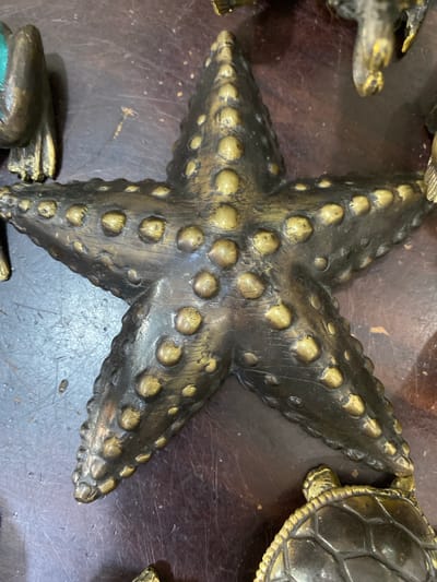 A high-angle, close-up shot depicts a dark metallic starfish, likely a decorative object, lying on a dark, possibly wooden, surface. The starfish has five arms and is textured with numerous raised, rounded bumps. Partial views of other dark metallic objects, including what appears to be a turtle figure, are visible near the edges of the frame. The environment suggests an indoor setting, possibly a shop or display area. The image does not contain any people. The location in Nusa Dua, Indonesia, is not evident from the image.