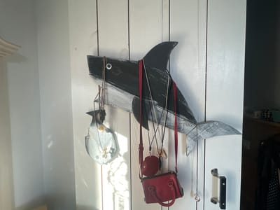 A wooden shark decoration is mounted on a white wooden door, serving as a holder for various accessories. The shark is painted black and white, with a grey tail and fins. Several necklaces with pendants hang from the shark's body and fins. Two small red handbags, a round red coin purse, and a seashell are also suspended from the shark. The scene is illuminated by sunlight streaming in from the left, casting shadows and reflections on the wall and door. The door has a black and silver handle on the right side. The overall impression is one of a casual, home setting.