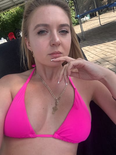 A young woman with blonde hair and light skin is lying down outdoors, wearing a bright pink bikini and a gold chain with a cross pendant. Her eyes are open and looking towards the camera, and her hand is resting near her chin. The background suggests a sunny day, with a tiled patio and greenery visible. The overall impression is one of relaxation and leisure in a backyard setting.