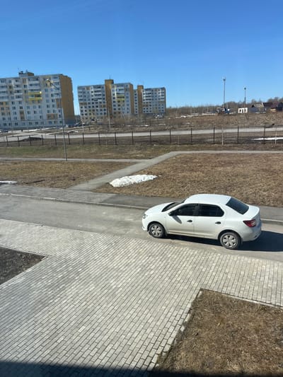 The image shows an aerial view of an urban scene in Berezniki, Russia. In the foreground, a white sedan is parked on an asphalt road adjacent to a brick-paved area. Patches of melting snow are visible on the grassy areas along the roadside. Further in the background, there are several multi-story apartment buildings. A chain-link fence runs along the road. The sky is blue and clear.
