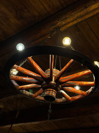 A wagon wheel chandelier hangs from a wooden ceiling. The wagon wheel is painted red with yellow and green decorative elements on its spokes. Several light bulbs are attached to the wheel, illuminating the scene. The ceiling is made of dark wooden planks. The image appears to be taken indoors, possibly in a restaurant or tavern in Bansko, Bulgaria, given the rustic aesthetic often found in the region.