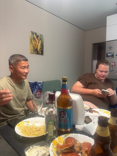 In an indoor setting, likely a dining area or kitchen, two individuals are present at a table laden with food and drinks. On the left, an adult male of East Asian descent, with short, graying hair, is seated and looking to his right, with his left hand gesturing. He is wearing a light olive-green t-shirt. To his right, a partially visible clear glass with a liquid and a small, ornate shot glass are on the table. A white plate with spaghetti-like noodles is in front of the man. A blue and white plastic bag with Cyrillic text is behind him.

On the right side of the frame, an adult female with light skin and brown hair pulled back is seated, looking down at her mobile phone, which she is holding with both hands. She is wearing a brown, short-sleeved top. A white takeout container sits on the table between her and the man.

The table also displays several bottles of alcohol, including a prominent bottle of Löwenbräu beer with a blue and gold label. Other bottles, some appearing to be beer and one a dark glass bottle with a red label, are also visible. Plates with food, including what appears to be fried fish and some vegetables, are arranged on the table. A small container of white dip is in the foreground.

The background reveals a wall with a framed artwork of stylized yellow and green leaves. Further back, a section of a refrigerator is visible, adorned with various magnets and a postcard featuring a landscape scene with Cyrillic text, possibly identifying a location within or near Novorossiysk, Russia. The overall lighting suggests an indoor environment, likely during daylight hours.