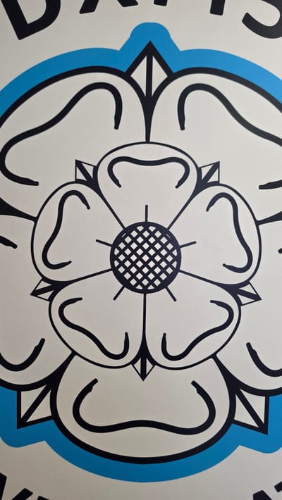 The image depicts a stylized Tudor rose, a traditional heraldic emblem of England. It is a white rose with black outlines, set against a light blue background. The center of the rose features a geometric pattern resembling a woven grid. The rose is likely part of a larger design or display, as only portions of it are visible. The Tudor rose emblem is used in various contexts, including civic symbols in the United Kingdom. The city of Bolton, United Kingdom is named because the specific location is not visually discernible in the picture.