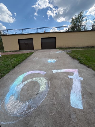 A ground-level perspective shows a concrete path with chalk drawings on it. The drawings include the numerals "6," "7," and smaller circular shapes. To the left of the path is a two-story building with a yellow brick facade, dark-colored garage doors, and a decorative black railing along the top of the building. Adjacent to the path is grass. The sky is visible with clouds. The location appears to be outdoors, possibly in Овсянка, Russia.