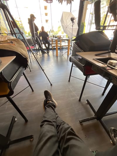 The image depicts an indoor scene within a modern establishment in Kafr Sousa Municipality, Syria, likely a cafe or restaurant, where a media production is underway. The perspective is from a seated individual, whose legs, clad in black trousers and black sneakers, extend into the foreground. To the left, a dark-colored jacket or bag rests on a table or chair. To the right, a black-framed table with a light-colored top features a recessed area containing small metallic objects, possibly production tools. Black tripod legs are visible on the dark tiled floor between the viewer and the central activity.
In the midground, illuminated by natural light entering through large windows, a man in a dark hoodie and cap stands behind a camera setup on a tripod. He faces another man seated at a light-colored wooden table. The seated man, dressed in dark attire, gestures with his hand near his face, with food items visible on the table before him. To his right, a large white photographic umbrella (softbox) is positioned, indicating artificial lighting for the production. Additional chairs and tables are arranged around the space, and a large black equipment case rests on a chair on the right side of the frame. Through the expansive windows in the background, outdoor trees and multi-story buildings are visible under daylight conditions. Decorative banners, one of which appears to read "SYRIA," hang from the ceiling.