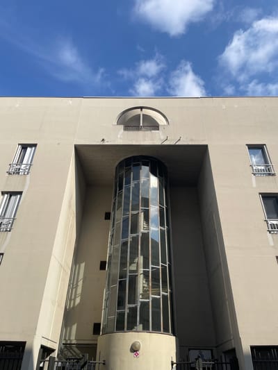 The image depicts the upper and mid-sections of a large, light beige-colored modernist building in Villeurbanne, France, under a clear blue sky with scattered white cumulus clouds. The building's facade features a prominent central, multi-story, cylindrical structure composed of numerous rectangular glass panes set within a light frame, indicating a vertical circulation shaft, possibly an elevator or stairwell. This cylindrical element is recessed within a large, arch-shaped opening in the facade. Above this arch, centered at the building's top, is a smaller semi-circular archway revealing a balcony with a dark railing.

To the left and right of the central features, the building presents multiple rectangular, vertically oriented windows, each fitted with a minimalist metal Juliet balcony or railing. Water streaks or stains are visible below some windows on the left side. The glass of the central cylinder reflects the blue sky and clouds, along with internal structures. At the base of the cylindrical structure, a circular light fixture is visible, alongside a small red circular sticker or emblem. Dark metal railings are present at the very bottom of the frame, hinting at a ground-level barrier. Strong, diagonal shadows are cast on parts of the building, indicating direct sunlight from the right. A small, indistinct poster featuring a person's image is barely discernible behind the lower right railing. No individuals are clearly visible or identifiable in the scene.