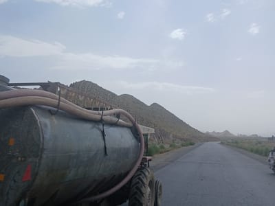 The image depicts a road scene in Qilla Saifullah, Pakistan. In the foreground, a large, cylindrical water tank is mounted on a tractor. The tank is grey and appears to be made of metal, with a prominent red triangular reflector on its side. A ribbed hose snakes across the top of the tank. The tractor's large rear tires and smaller front tires are visible.

The road stretches into the distance, bordered by dry, arid hills on the left and sparser vegetation on the right. The landscape is mountainous and rocky, typical of a desert or semi-arid region. In the far distance, more subdued hills are visible under a hazy sky.

The sky is overcast with scattered clouds, suggesting either early morning or late afternoon, or perhaps a generally cloudy day. The overall atmosphere is one of a dry, possibly hot, climate. The presence of the water tank suggests a need for water transport, perhaps for agricultural or domestic purposes in this arid environment.

On the right side of the road, a person on a motorcycle can be seen in the distance, hinting at the presence of ongoing activity or travel. There is no visible text in the image. The scene conveys a sense of travel and the practicalities of life in a challenging geographical setting.