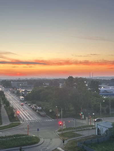 The image shows a sunset over the city of Kemerovo, Russia. The sky is filled with vibrant orange and pink clouds, with a gradient of blue towards the top. In the foreground, a street intersection is visible with cars and trucks on the road. Traffic lights are red, indicating that vehicles should stop. Pedestrians are crossing the street. Trees and buildings line the street, and in the distance, industrial smokestacks can be seen. The overall impression is a busy urban environment bathed in the warm glow of the setting sun.