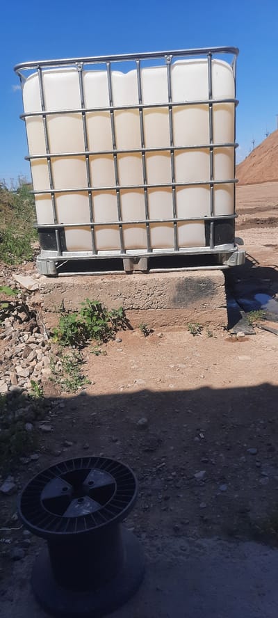 The image depicts an outdoor scene on a sunny day, with a bright blue sky and scattered clouds. In the foreground, a black plastic cable spool rests on a gravelly, sandy surface. The ground is uneven, with visible stones and debris.

Dominating the middle ground is a large, white IBC (Intermediate Bulk Container) tote. This container is encased in a silver metal cage and sits on a concrete platform. The tote is partially filled with a light-colored liquid. The concrete platform appears weathered and is surrounded by more gravel and some sparse green vegetation.

In the background, to the right, a large pile of reddish-brown earth or sand rises up. The overall environment suggests an industrial or construction site, possibly in a rural or undeveloped area. There are no people visible in the image. The location is identified as Zhasagan, Kazakhstan.