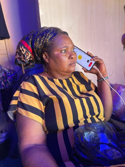 A middle-aged Black woman is lying on her side, holding a black smartphone to her right ear with her right hand. The phone screen is displaying a video call interface. Her head is wrapped in a patterned scarf. She is wearing a yellow and black striped shirt. A charging cable is connected to her phone. She wears a wristband. She is lying on a surface partially covered with black plastic bags. The background appears to be an interior space with a light-colored wooden surface. The location is Samaru, Nigeria.