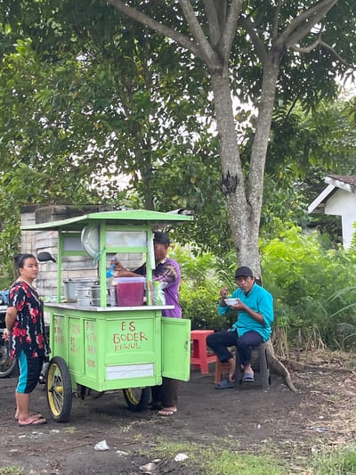 In Jati Alun Alun, East Java, an outdoor scene unfolds at a food cart. A woman stands to the left of a bright green cart, which displays text in red that reads "ES GODER KAWUL". She is wearing a black shirt with a floral pattern and blue shorts. A man in a purple shirt is behind the cart, holding a blue ladle and dispensing a pink liquid into a cup. To the right, a man in a teal shirt and black cap sits on a stool, eating from a white bowl. He is positioned near a large tree trunk. The cart is laden with various metal containers. The ground is dirt and some patchy grass. In the background, lush green foliage and a white building with a prominent roof are visible.