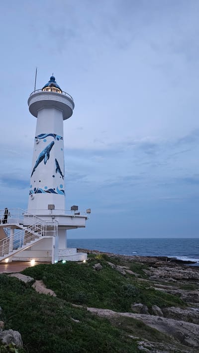 The image shows a white lighthouse decorated with blue whales and wave patterns. The lighthouse is positioned on a coastal cliff with lush green vegetation and rocky formations. In the background is the ocean, leading to a cloudy sky. The overall view suggests a seaside location. This is the GanJeolGot Lighthouse in Ulsan, South Korea.