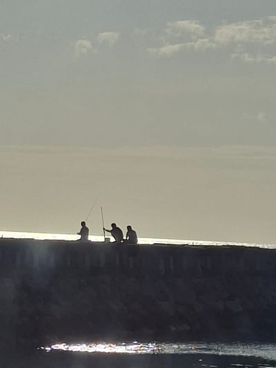 Three silhouetted figures are fishing from a stone breakwater under a hazy sky with scattered clouds. The figures are positioned at varying distances apart, with the leftmost figure holding a fishing rod upright. The middle figure is bent over, also holding a fishing rod. The rightmost figure is seated. The breakwater is dark and appears to be made of rough stones. Below the breakwater, the water reflects the light, creating a shimmering effect. The scene is captured at a distance, with the focus on the silhouettes against the bright, overcast sky. The location is identified as Nusa Penida, Indonesia.