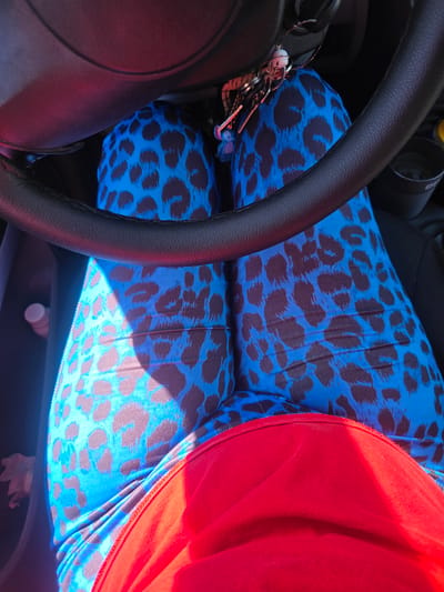 The image shows a person's legs and lap inside a vehicle, with a steering wheel visible in the foreground. The person is wearing bright blue leggings with a dark brown leopard print pattern and a red garment, possibly a skirt or dress, is draped over their lap. Keys with several charms hang from the steering column. The environment appears to be a car interior, with a cup holder visible to the right. The lighting suggests it is daytime. The image does not contain any information that would place it in Kostinbrod, Bulgaria.