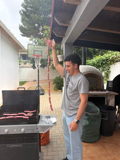 A man stands outdoors near a grill, holding a string of uncooked sausages in his extended hand. Some sausages are already cooking on the grill. A basketball hoop stands nearby in a yard surrounded by buildings and greenery. The location appears to be Montgó, Spain.