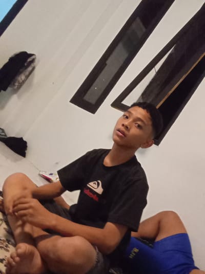 The image shows a young person sitting on a patterned mat or rug in what appears to be an indoor setting. The person is wearing a black t-shirt with a logo that includes a stylized wave and the word "Quiksilver" in red text. They are also wearing blue shorts. Their legs are crossed, and their hands are resting on their lap. The person is looking towards the camera with a neutral expression.

Behind the person, the wall is painted white, and there are two dark-framed windows with a reflective surface, suggesting they might be glass or polished wood. The windows are positioned diagonally on the wall. To the left of the frame, some dark clothing is visible, draped over a surface.

The lighting suggests it could be evening or indoors with artificial light, as there are no strong shadows indicative of direct sunlight. The overall scene is casual and appears to be a candid shot. The provided location context is Tibojong, Indonesia. There is also some faint, unidentifiable text on the blue shorts.