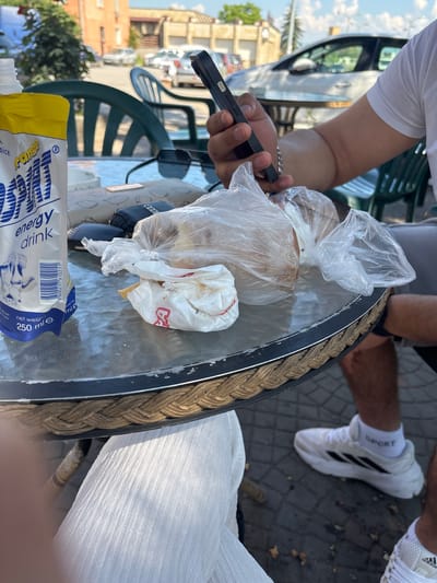 This image captures a close-up view of a glass-topped table with various items on it. In the foreground, a person's hand holds a dark-colored smartphone. To the left, a yellow and white pouch, likely for an energy drink, is visible, featuring blue text and graphics including a barcode and nutritional information. A white plastic bag containing what appears to be bread or pastries is situated in the center of the table. Scattered around are a pair of dark sunglasses, a wallet with a patterned design, and a black car key. The table is surrounded by green plastic chairs. In the background, a white car and other vehicles are parked, suggesting an outdoor setting, possibly a cafe or restaurant, given the table and chairs. The environment appears to be urban, with paved ground and buildings in the distance. This scene is located in Elin Pelin, Bulgaria, evidenced by the presence of the Elin Pelin municipality emblem on the energy drink pouch.