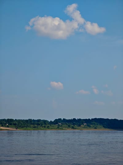 The image depicts a wide, calm river flowing in front of a densely forested embankment under a bright blue sky with scattered white clouds. On the embankment, which appears to be the outskirts of Ust'-Kem', Russia, there are a few scattered buildings and some vehicles parked on a dirt track. The water surface is rippled, reflecting the light of the sky. The overall scene is serene and natural, with a hint of human presence on the distant shore.