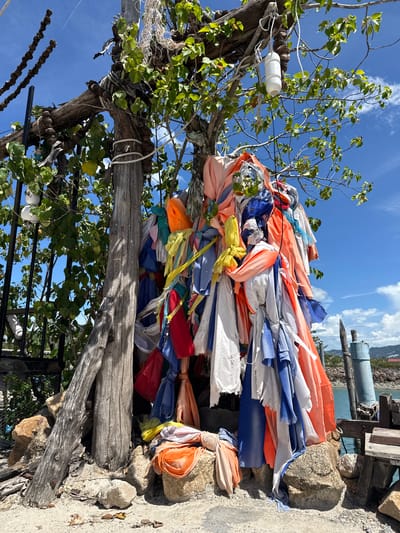 In Baan Plai Laem, Thailand, a rustic wooden structure, seemingly a shrine or monument, is adorned with numerous colorful fabric strips tied in knots. These strips, in shades of orange, blue, white, pink, and yellow, hang from branches and a central wooden pole. The structure is situated outdoors, with a clear blue sky and scattered clouds visible above. Green foliage from a tree with broad leaves partially obscures the shrine and the sky. The base of the structure is surrounded by rocks and sand, with a hint of the ocean and a blue structure in the background, suggesting a coastal location. A metal fence with vertical bars is visible to the left of the wooden structure.