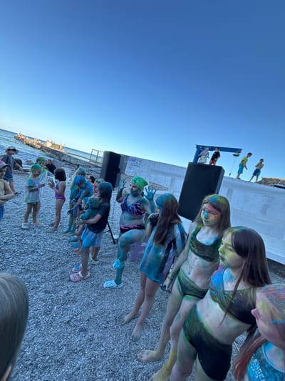 The image captures an outdoor gathering where people, mostly young individuals, are participating in a color powder event. The subjects are positioned along a rocky shoreline, with the sea and a clear blue sky as the backdrop. The individuals are covered in various colors of powder, suggesting a festival or celebration of some sort, possibly a Holi-like event.
The scene is set during the day, indicated by the bright sunlight and the clear blue sky. The location appears to be a public space by the sea, with a concrete structure and a large black speaker on a platform, suggesting a planned event with music or announcements. People are dressed casually, in swimsuits or light clothing, suitable for warm weather. Some are standing in a line, while others are seated or interacting.
Notable details include the vibrant colors of the powder on the subjects' skin and clothing, creating a festive and playful atmosphere. The expressions on some faces suggest engagement and enjoyment. The presence of the sea and the rocky ground are strong location cues.
There is some visible text on the clothing of a person in the upper right quadrant of the image. The text, partially obscured, appears to read "ANGEZ". The overall mood is lively and spirited.