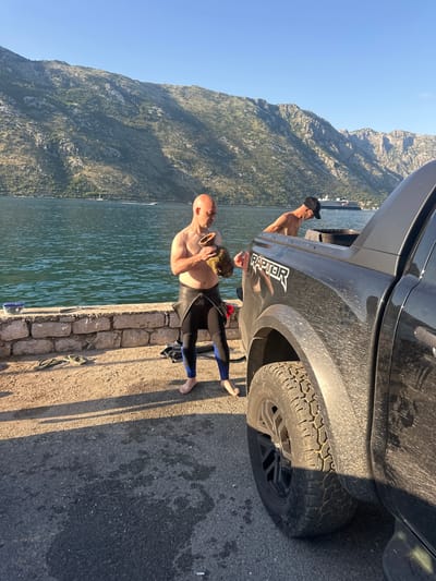 The image depicts a coastal scene in Prčanj, Montenegro, featuring two men and a dark pickup truck by a body of water with mountains in the background under a clear sky.
In the left-center, a bald, shirtless Caucasian male with a light tan is standing barefoot on a paved surface. He is wearing the lower half of a black and blue wetsuit, with the top half pulled down to his waist. He is holding two irregular, organic-looking brown objects in his hands, appearing to examine them. One object, larger, is in his left hand, and a smaller one in his right. His gaze is directed downwards at these objects.
To the right, partially obscured by a dark Ford Raptor pickup truck, a second leaner Caucasian male, wearing a black baseball cap and bare-chested, is bent over. He appears to be interacting with the bed of the truck, possibly retrieving or stowing items. The truck is dark-colored, visibly dusty, and features "RAPTOR" branding on its side. A robust tire with an aggressive tread pattern is visible on the front passenger side.
The ground consists of a paved surface (asphalt or concrete) in the foreground, transitioning to a stone wall or embankment directly adjacent to the water. A pair of yellow-green flip-flops and a blue plastic container with a clear lid are resting on the paved ground near the wall.
The body of water in the background is calm with visible ripples, reflecting the sunlight. Beyond the water, steep, rocky mountains covered with sparse vegetation rise prominently. A large cruise ship is visible in the mid-distance on the water, positioned to the left of the second man and the truck. Smaller boats, including a sailboat, are also discernible further in the distance. The lighting suggests either morning or late afternoon, with strong, directional sunlight creating distinct shadows on the ground.